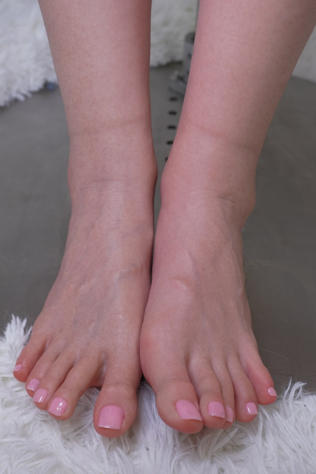 Close-up image of a pair of feet with a light pink nail polish, resting on a soft surface. The feet are positioned together, showcasing details of the toes and skin texture.